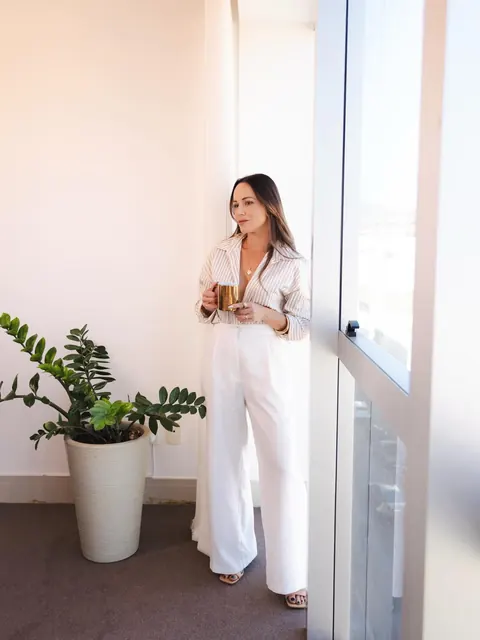 De macacão branco, ela segura uma xícara perto da janela clara, com uma planta ao lado