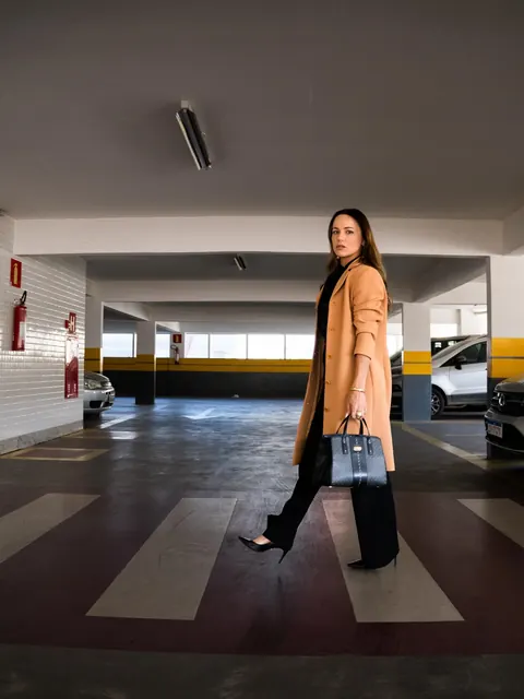 De sobretudo caramelo e bolsa na mão, ela atravessa a faixa de pedestres de um estacionamento