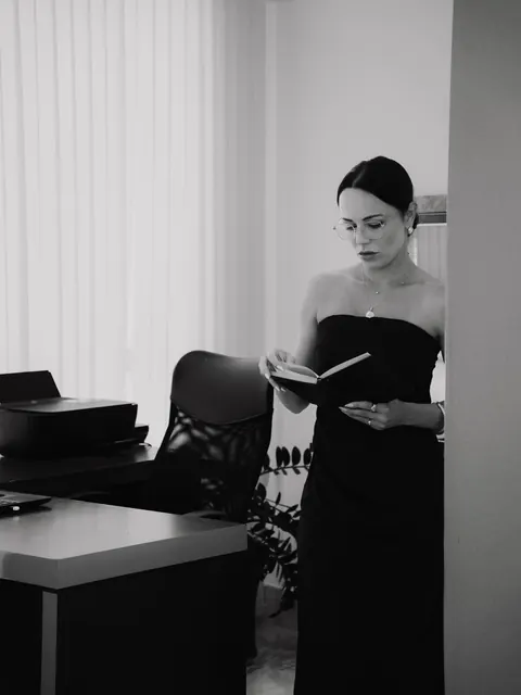 Em preto e branco, ela lê um caderno de pé, de vestido tomara que caia, ao lado da mesa do escritório