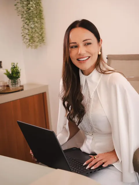 Ela sorri aberto olhando para o lado, com as mãos no notebook