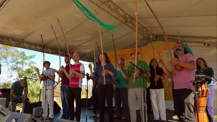 Capa do vídeo: músicos de capoeira cantam e tocam berimbau sob uma tenda, com bandeirinhas ao fundo
