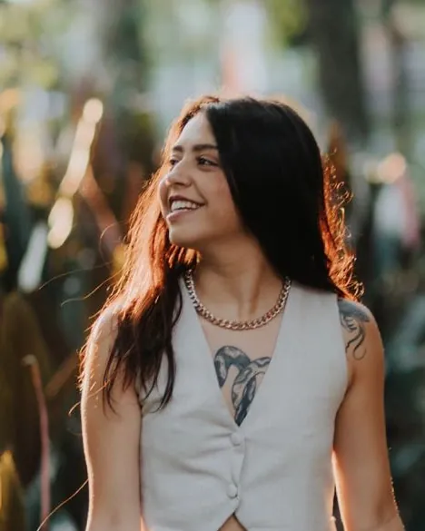 Lelet Barbosa sorrindo de olhos fechados no jardim, com colete branco e colar de corrente